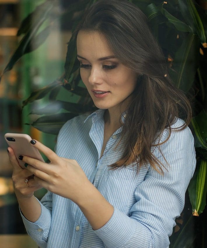 una mujer en un fondo colorido. tiene el pelo oscuro y lleva una camisa azul claro. Está mirando su teléfono